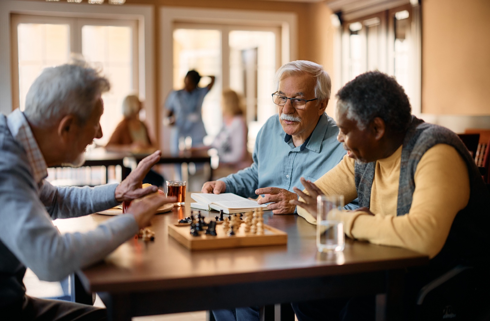 Seniors playing chess together while a senior spectator watches them with an open book in front of him.
