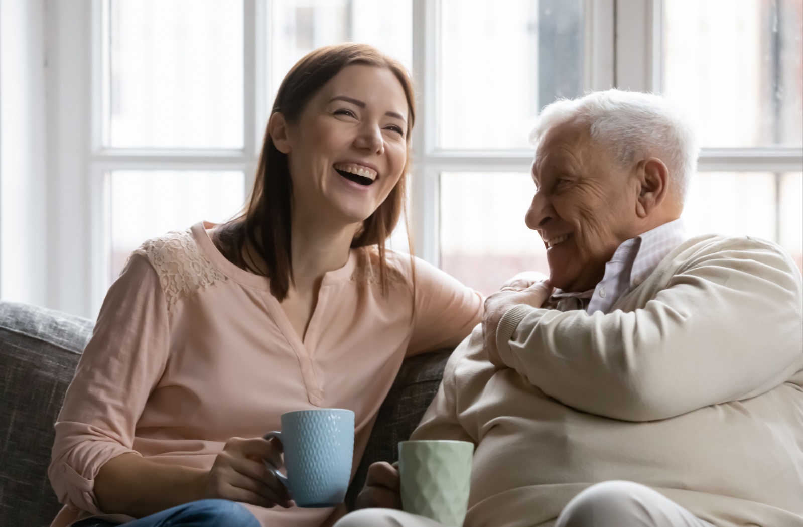 A woman and her senior father holding mugs and laughing during a visit in assisted living.