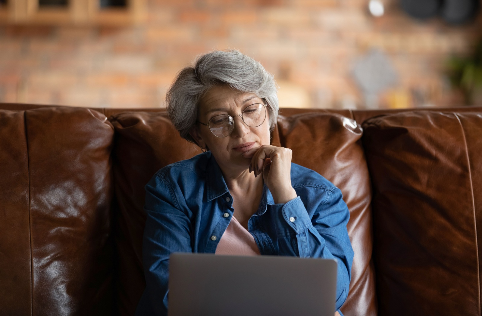 Thoughtful-looking senior woman using laptop while sitting on a leather couch.