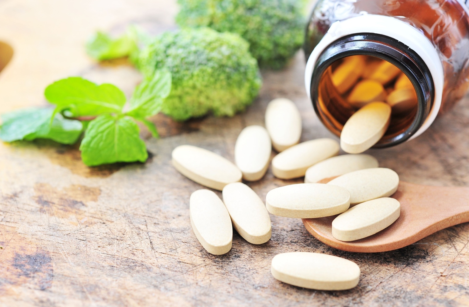 A bottle of vitamin C supplements lying on its side on a wooden table next to fresh vegetables.