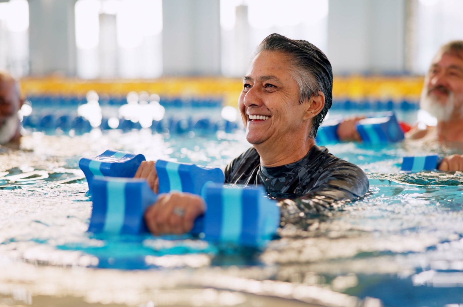 A smiling older adult with weights in both hands working out in an indoor pool.