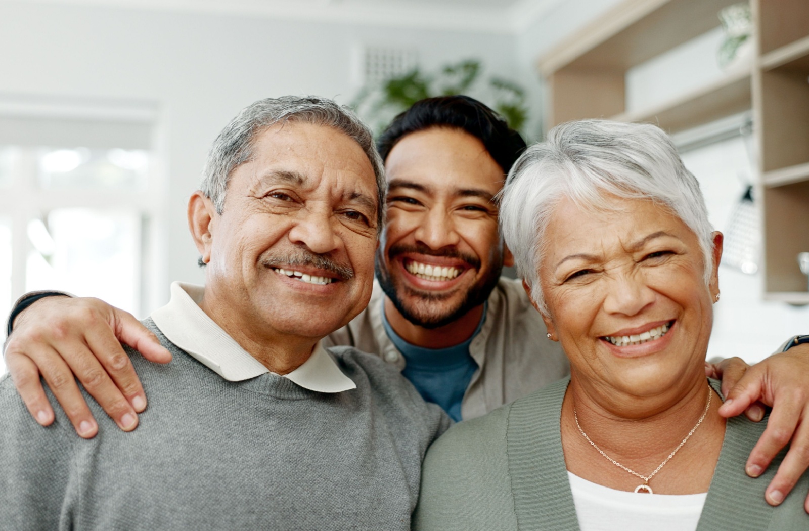 An adult child embraces their senior parents. All three people are smiling brightly.