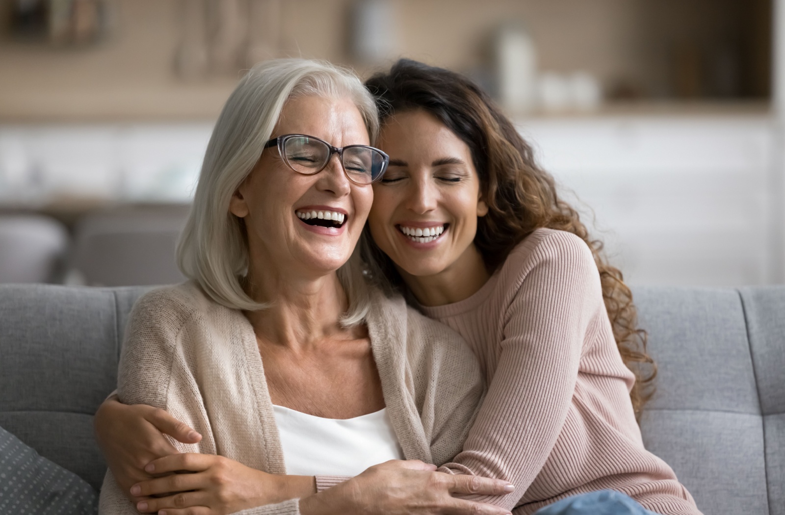 A person hugging their senior mother on the couch and laughing and smiling together.
