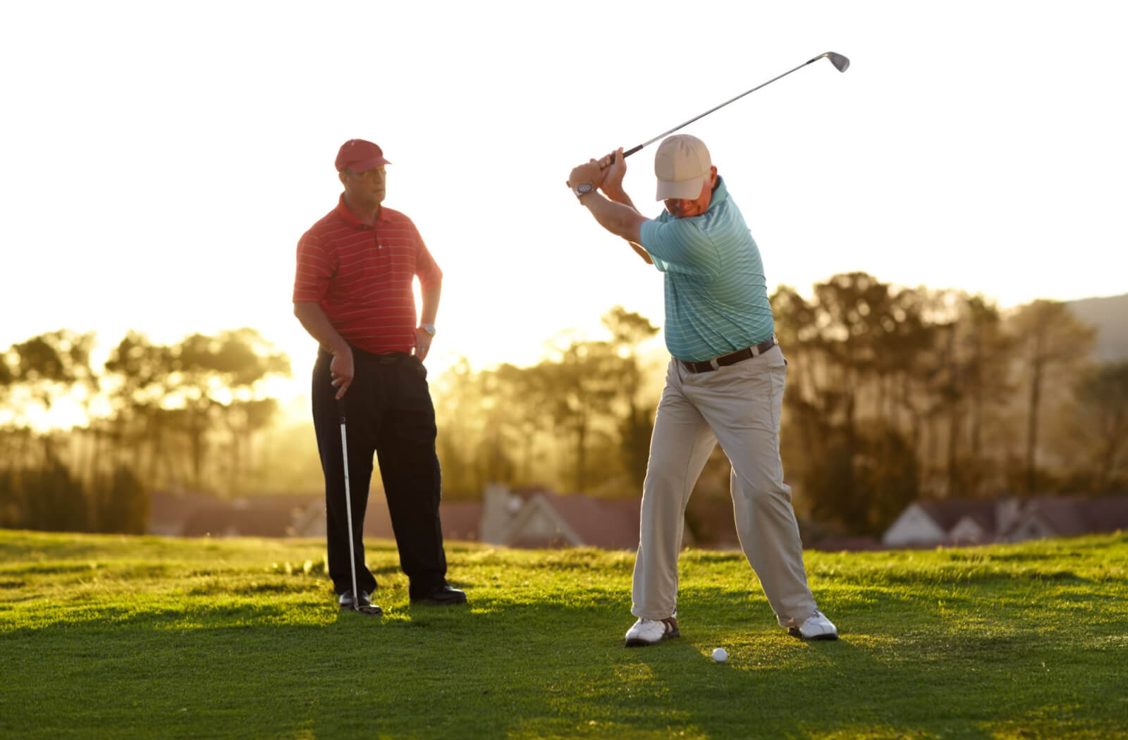 A wide shot of two older adults on the golf course mid-swing while the sun shines bright behind them.