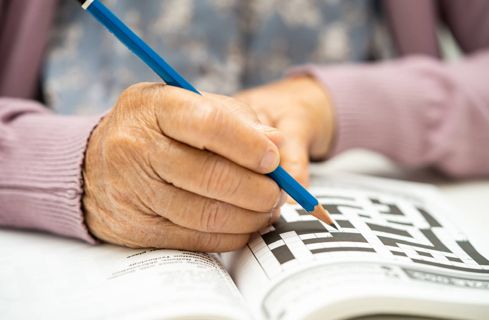 A close-up image of an older adult's hand holding a blue pencil while they work on a crossword puzzle to maintain their cognitive abilities
