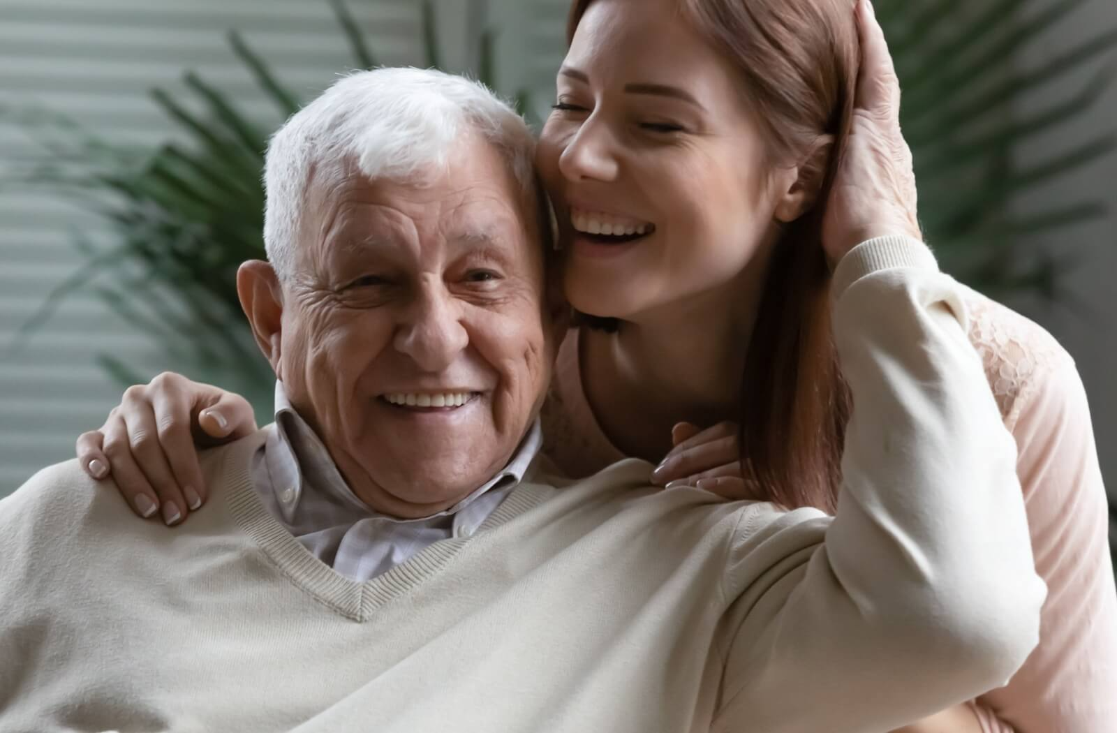 An adult child hugs their older parent from behind while they both laugh during a visit in assisted living.
