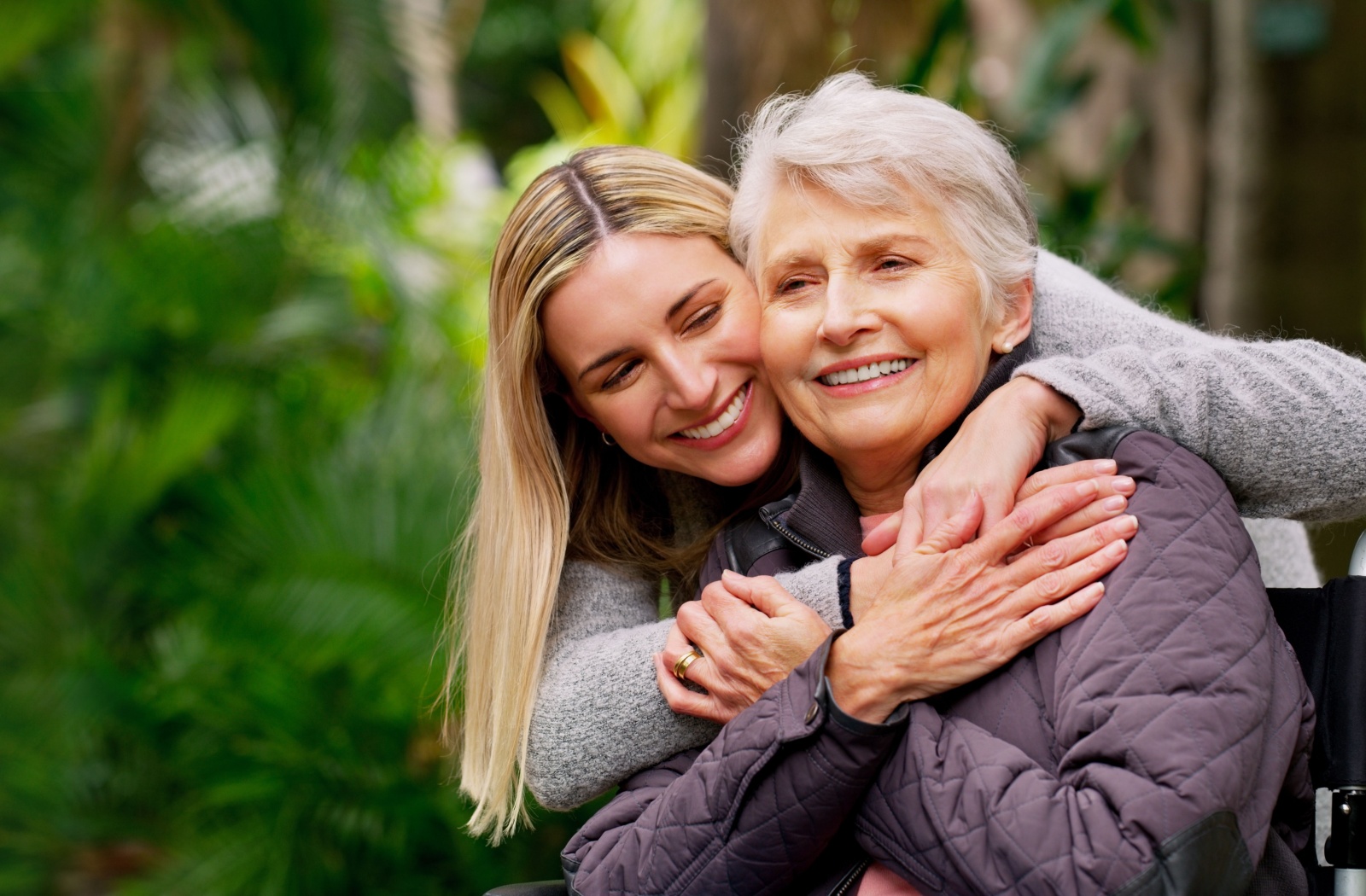 An older adult smiles while their adult child hugs them from behind while walking together outside.
