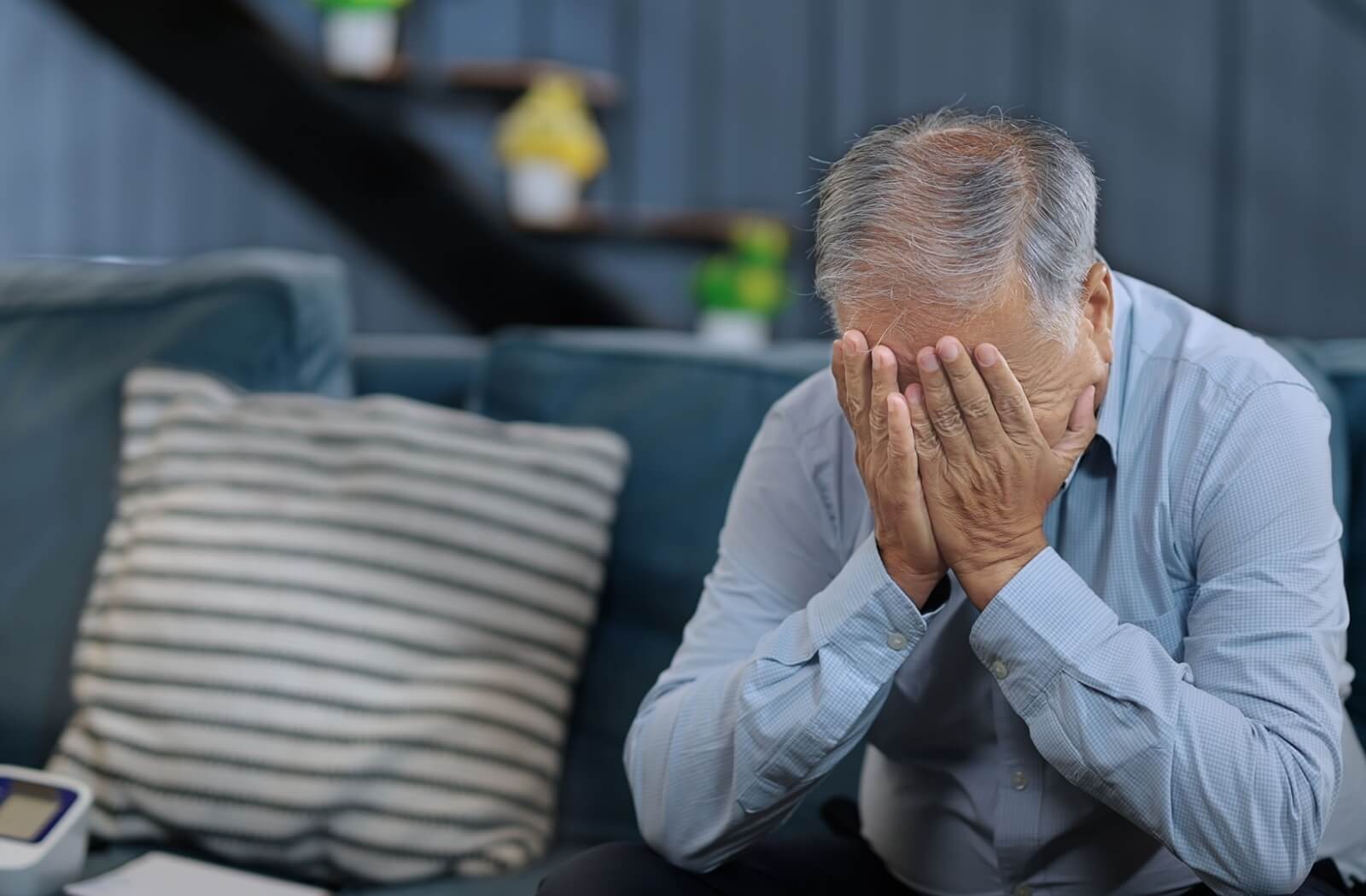An older adult sits on their couch and rubs their face in frustration due to their recent forgetfulness