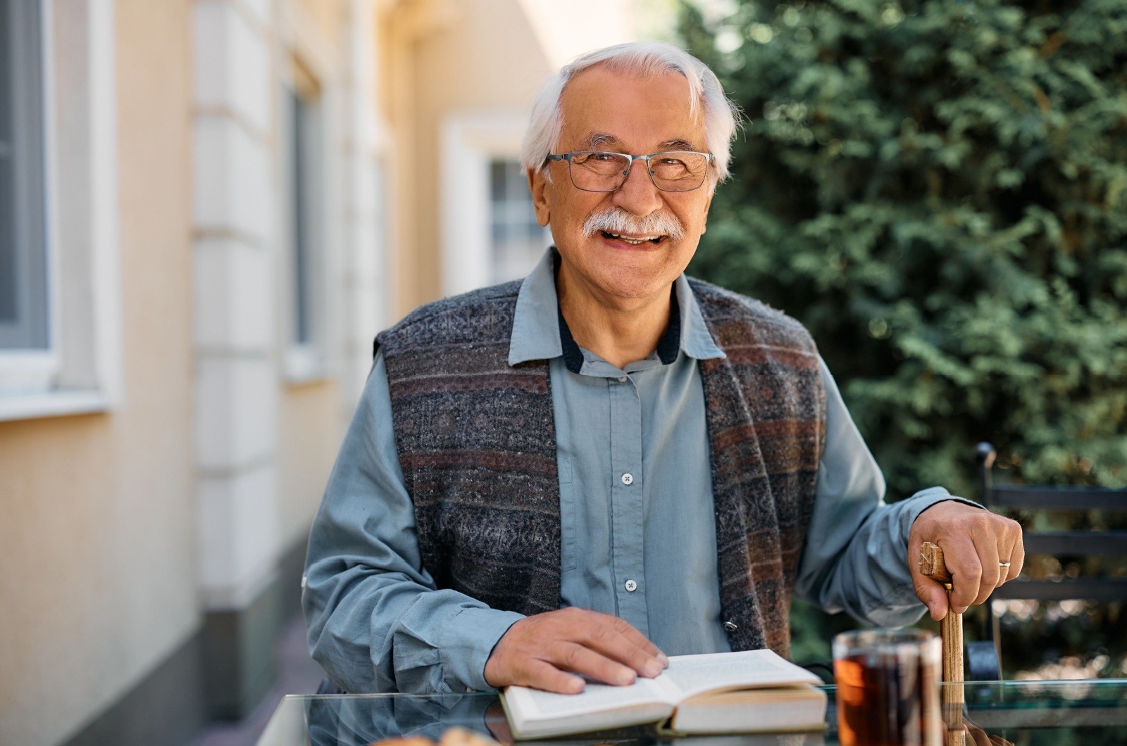 A smiling senior reads a book while sitting outside at a glass table.
