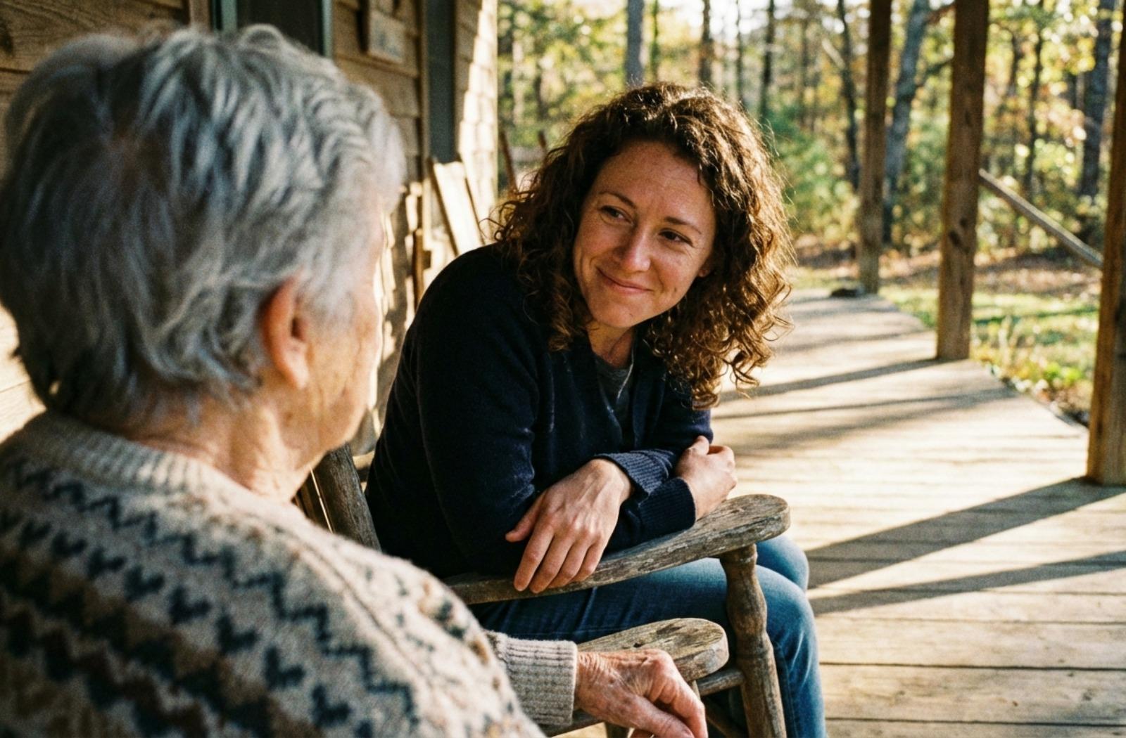 Younger adult sits on a porch, leaning forward with an empathetic expression while looking at an older person seated across from them.
